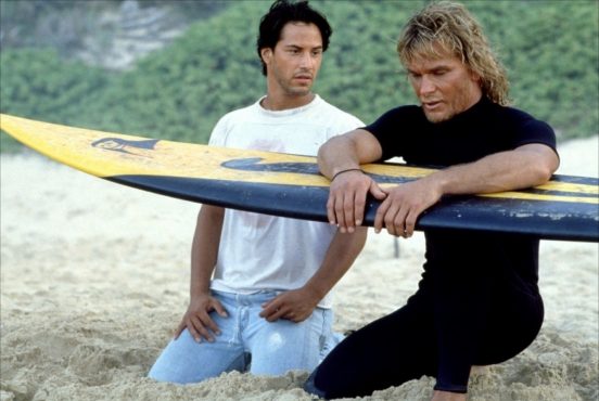 Cena do filme "Caçadores de Emoção" com Keanu Reeves e Patrick Swayze sentados na praia com uma prancha de surf