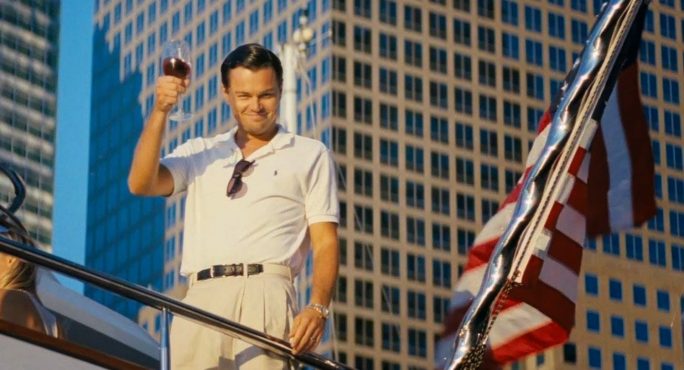 empreendedorismo no cinema com o filme "O Lobo de Wall Street". Na foto, cena do filme com o Leo Dicaprio levantando uma taça de vinho e uma bandeira do EUA ao lado.