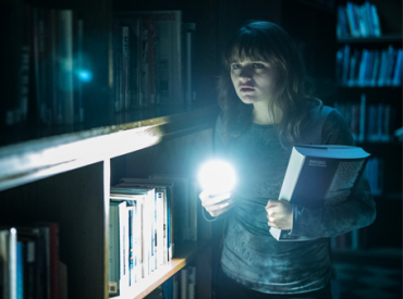 Cena do filme "Slender Man". Uma menina dentro de uma biblioteca escura, com um livro na mão e uma laterna na outra, observando o fundo de um corredor