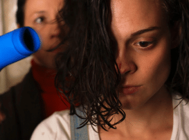 Foto divulgação do filme "Tesnota". Mulher jovem com o cabelo escondendo a metade do rosto, olhando para baixo e uma mulher mais velha atrás com um secador de cabelo