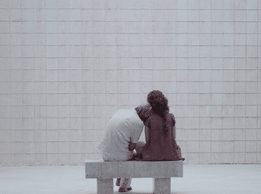 Foto de divulgação do filme "Unicórnio". Um home todo de branco com a cabeça deitada no ombro de uma mulher de vestido lilás, os dois sentados em um banco de pedra branco em um cômodo todo no azulejo branco.