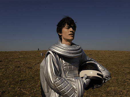 Filme Yonlu. Foto divulgação, jovem com roupa improvisada de astronauta onde o capacete é um modelo para motocicleta. Ele está em um campo aberto com gramado ressecado