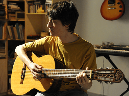 Foto divulgação do filme Yonlu. Jovem tocando violão em um quarto. com teclado ao fundo e uma prateleira cheia de livros e outros objetos.