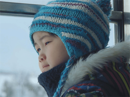 foto divulgação do filme "Takara a noite em que nadei". Menininho oriental com roupas para frio intenso, dentro de um transporte público com a cabeça encostada no vidro, observando a neve.