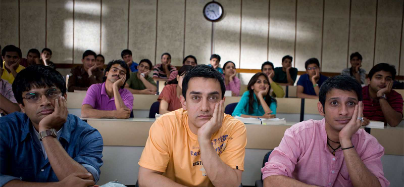 cena do filme indiano "três idiotas". jovens em sala de aula de uma universidade, todos com a mão no queixo. Sendo três na frente em destaque.